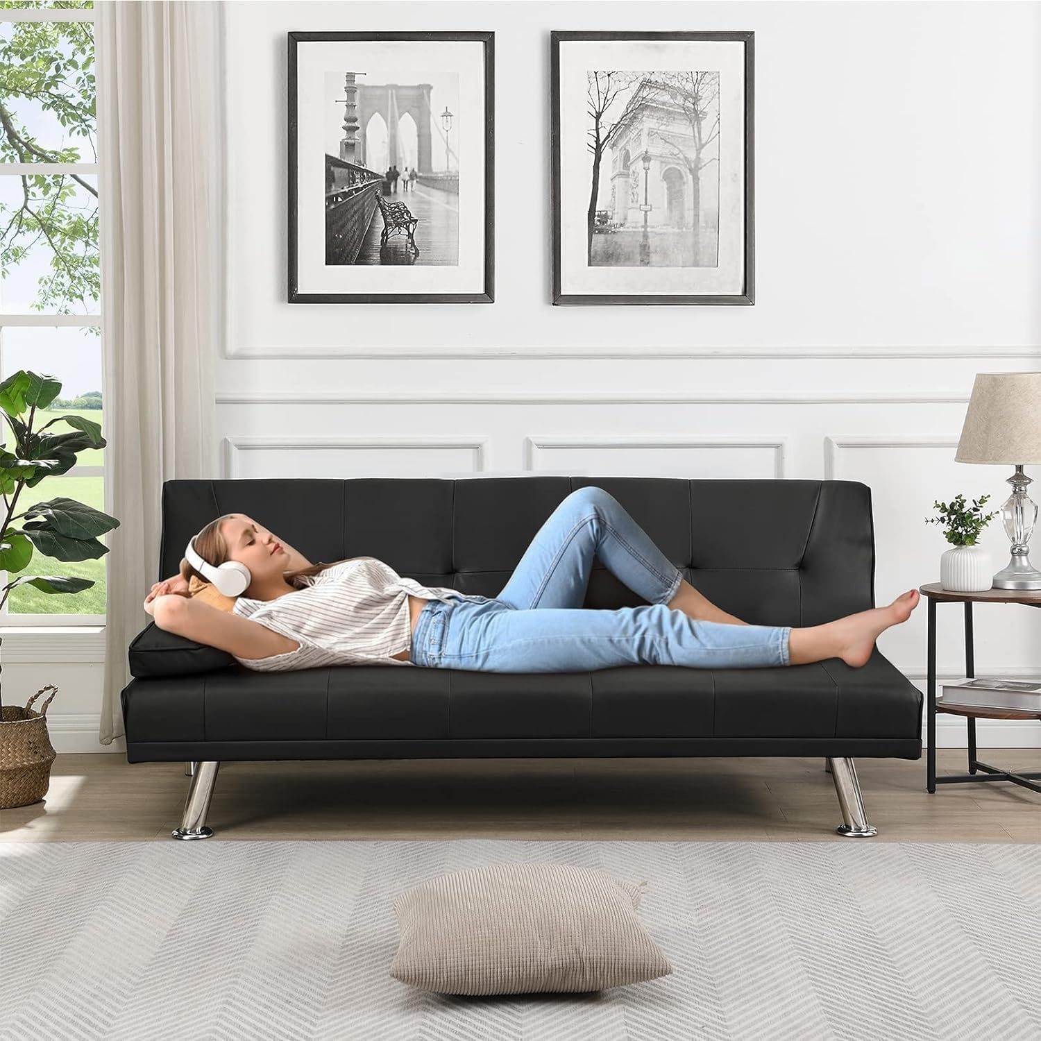 Couch Bed Futon Sleeper Sofa for Living Room | Black Faux Leather from AHF Depot - Black leather futon with center cupholders displayed in the couch position with a woman sleeping on it in a light-toned modern living room scene against a window sill. 