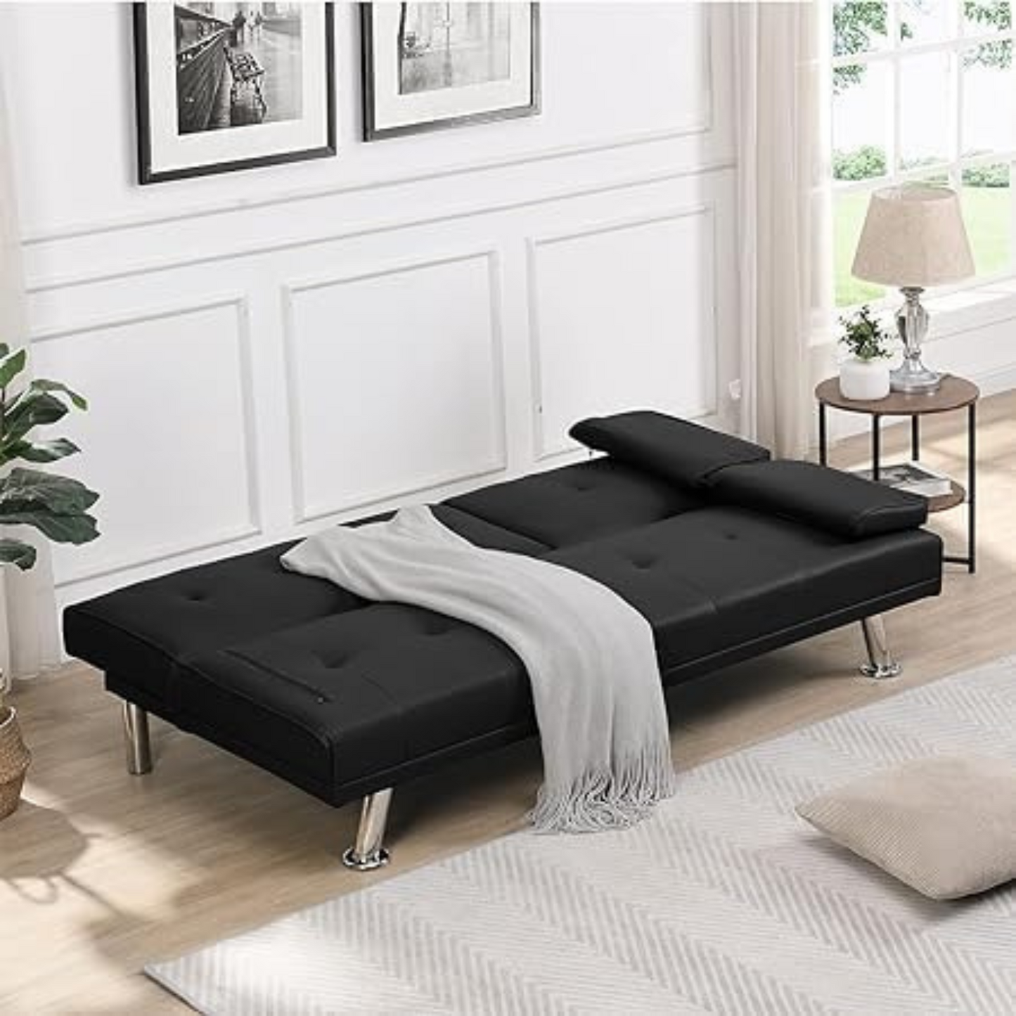 Couch Bed Futon Sleeper Sofa for Living Room | Black Faux Leather from AHF Depot - Black leather futon with center cupholders displayed in the bed position with a blanket on it in a white-washed modern living room scene. 